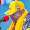 Man speaking into a red microphone, wearing a bright yellow cap and matching scarf at a public event. The cap shows the number '001' and he wears a patterned shirt with yellow accents.