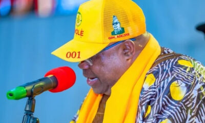 Man speaking into a red microphone, wearing a bright yellow cap and matching scarf at a public event. The cap shows the number '001' and he wears a patterned shirt with yellow accents.