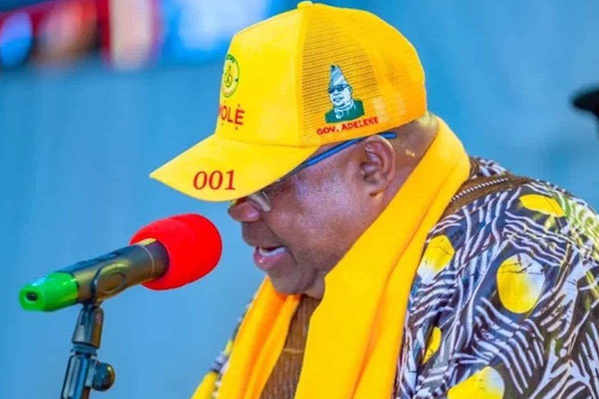 Man speaking into a red microphone, wearing a bright yellow cap and matching scarf at a public event. The cap shows the number '001' and he wears a patterned shirt with yellow accents.