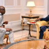 Two men sit on gold-upholstered chairs in a formal room, conversing near a side table with a lamp.