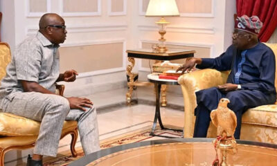 Two men sit on gold-upholstered chairs in a formal room, conversing near a side table with a lamp.