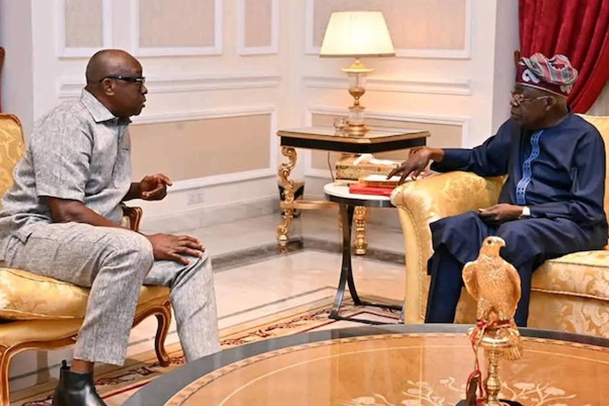 Two men sit on gold-upholstered chairs in a formal room, conversing near a side table with a lamp.