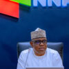 Man in white traditional attire and patterned cap sits at a desk with a microphone in front, in a room with an INEC backdrop behind him.