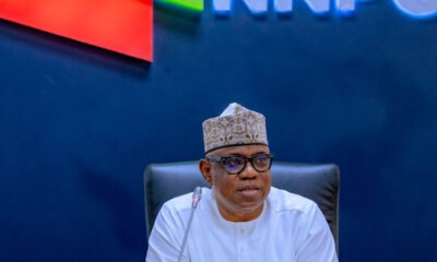 Man in white traditional attire and patterned cap sits at a desk with a microphone in front, in a room with an INEC backdrop behind him.