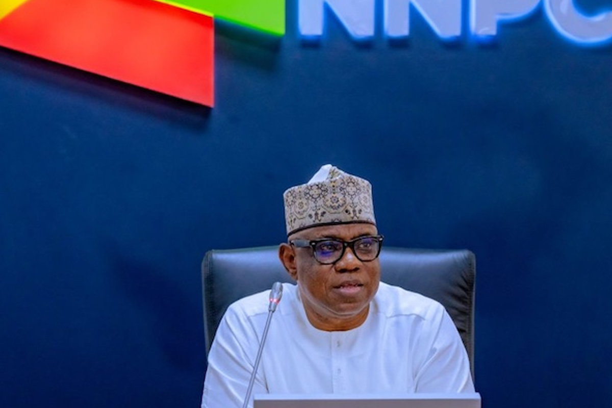 Man in white traditional attire and patterned cap sits at a desk with a microphone in front, in a room with an INEC backdrop behind him.