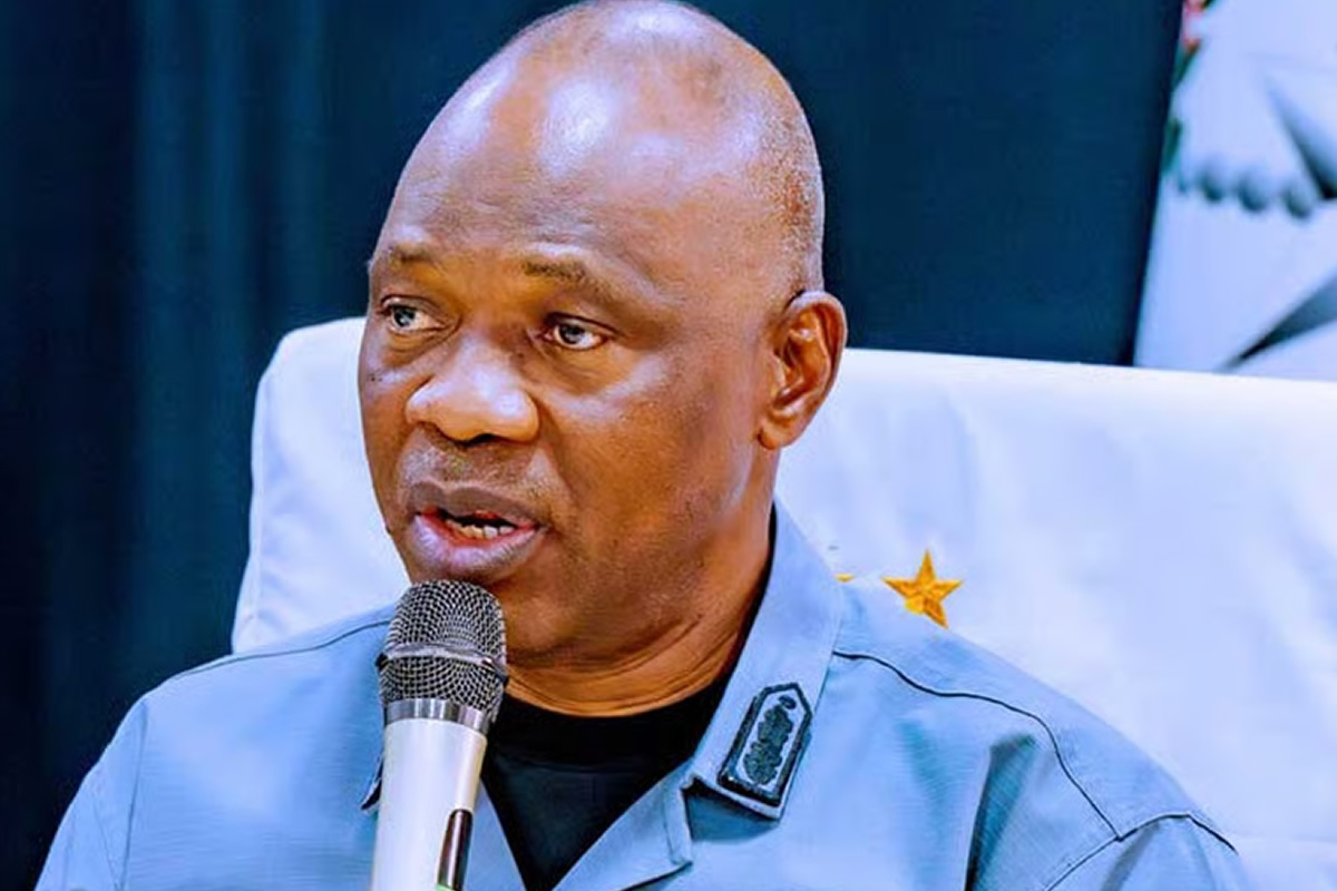 Man in uniform speaks into a handheld microphone at a public event, seated in a chair with a star insignia on the shoulder patch behind him