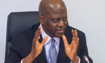 Man in a dark suit and light tie seated at a conference table, speaking with animated hand gestures toward an audience or camera.