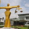 Golden statue of a muscular figure holding scales of justice in a public plaza outside a building