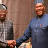Two smiling men in traditional West African attire shake hands indoors.