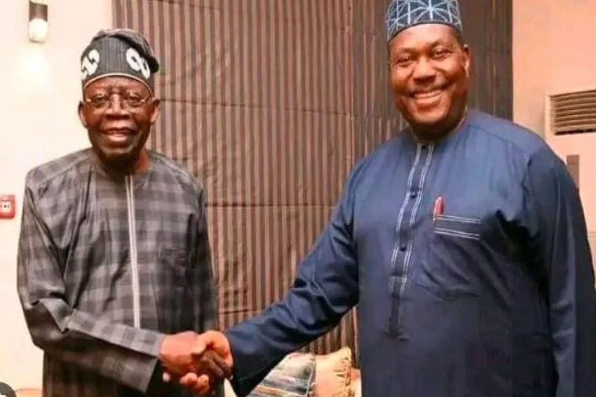 Two smiling men in traditional West African attire shake hands indoors.