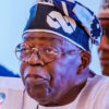 Elder Nigerian man speaking into a microphone, wearing glasses and a patterned headwrap with white attire at a formal event.