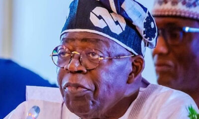 Elder Nigerian man speaking into a microphone, wearing glasses and a patterned headwrap with white attire at a formal event.
