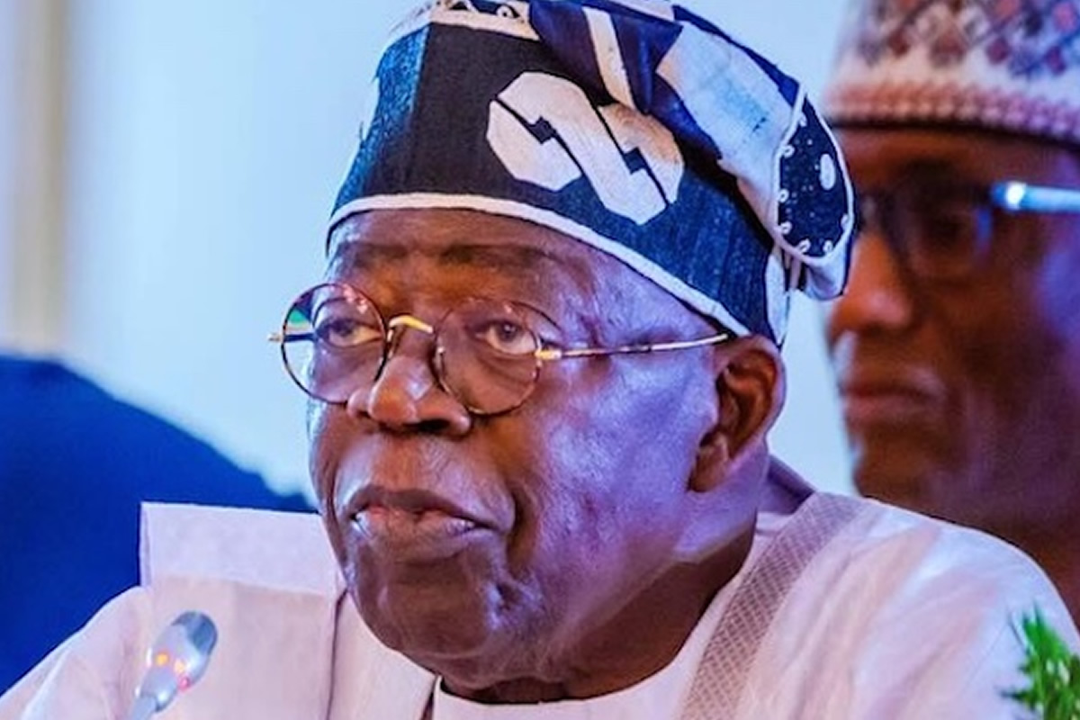 Elder Nigerian man speaking into a microphone, wearing glasses and a patterned headwrap with white attire at a formal event.