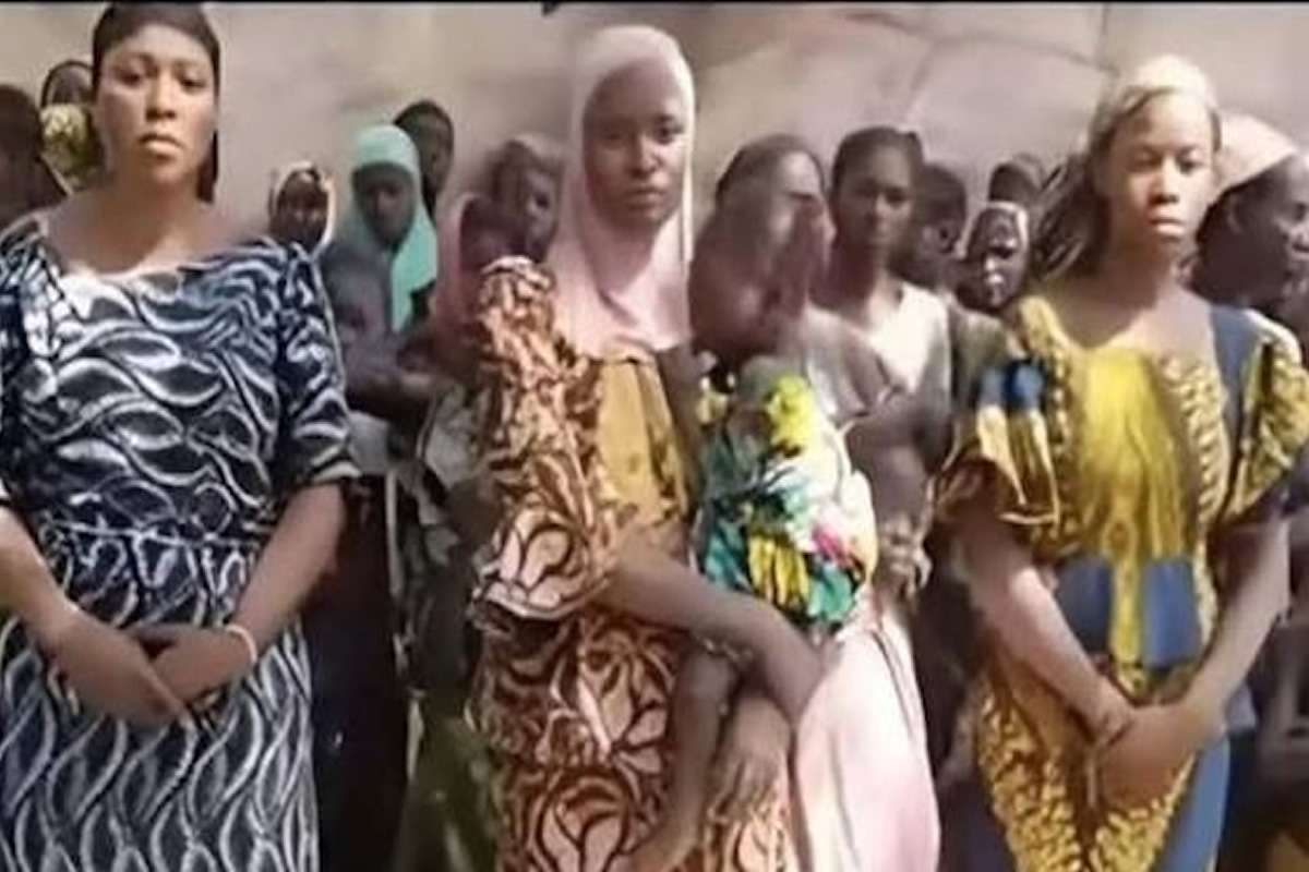 Group of women in colorful traditional dresses standing in a crowd, looking ahead at an event. Mention of a summer gathering or ceremony implied by attire.