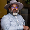 Portrait of a distinguished man wearing a gray hat, glasses, and a light blue striped shirt with a decorative necklace and pocket square.