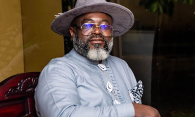 Portrait of a distinguished man wearing a gray hat, glasses, and a light blue striped shirt with a decorative necklace and pocket square.
