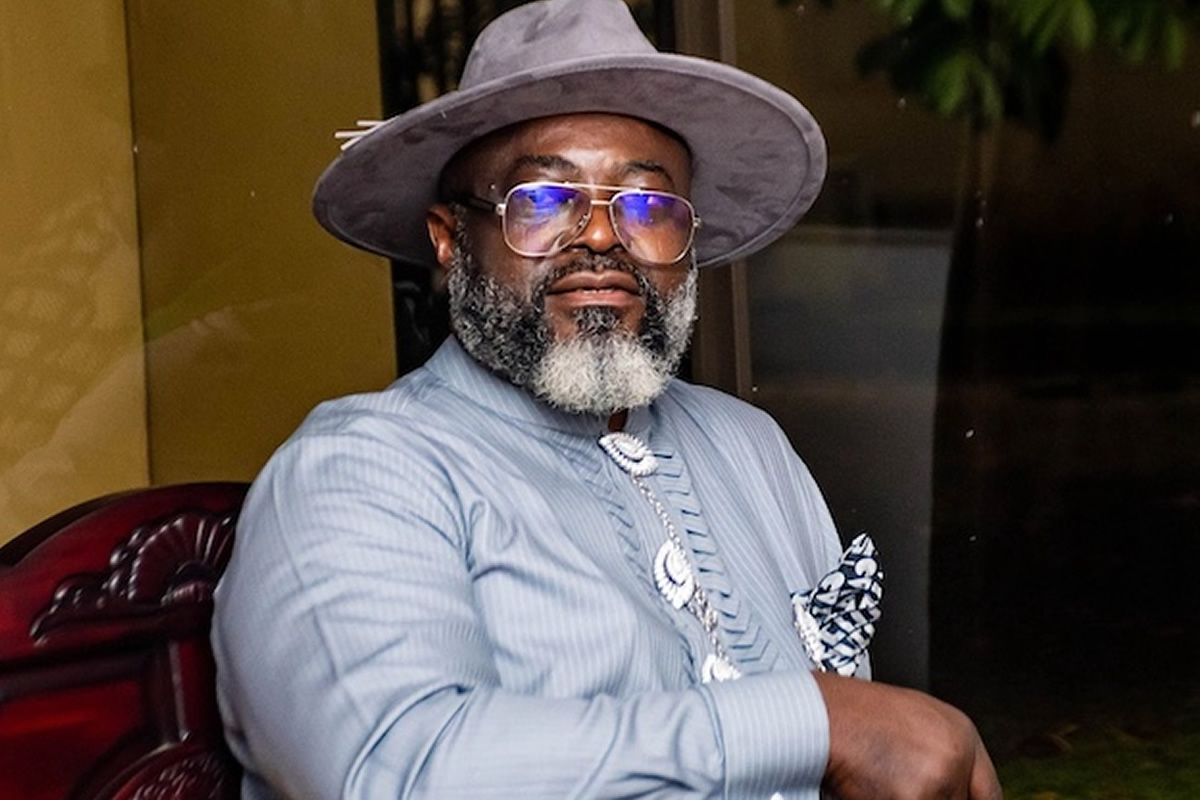 Portrait of a distinguished man wearing a gray hat, glasses, and a light blue striped shirt with a decorative necklace and pocket square.
