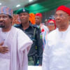 Two men in traditional attire walk side by side at a formal event, one with a hand on his chest beside security personnel.