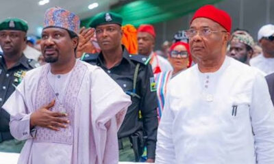 Two men in traditional attire walk side by side at a formal event, one with a hand on his chest beside security personnel.