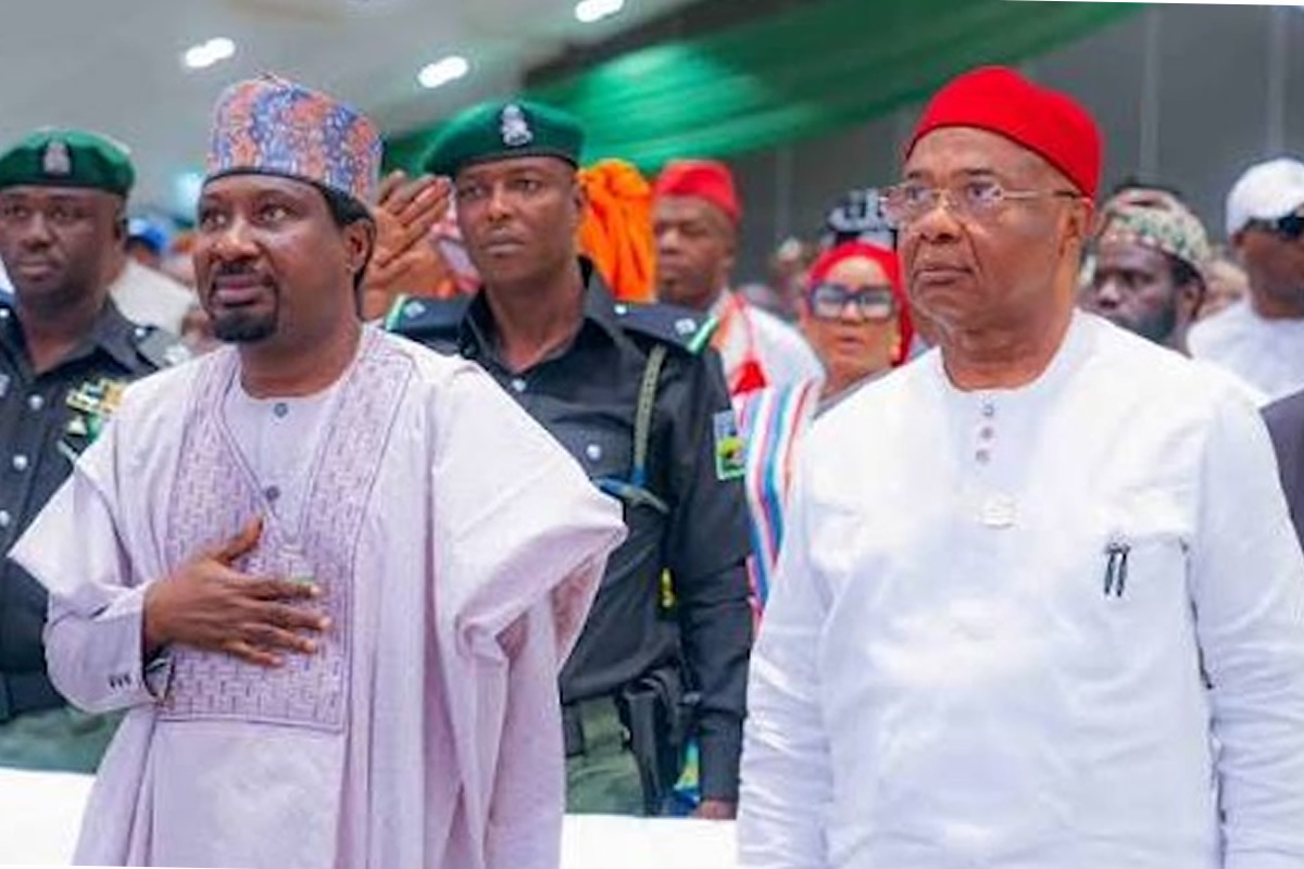Two men in traditional attire walk side by side at a formal event, one with a hand on his chest beside security personnel.